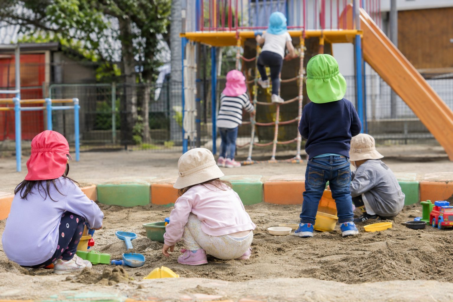 砂場でスコップを使って遊ぶ子どもたちと、奥のジャングルジムを登る子どもの後ろ姿